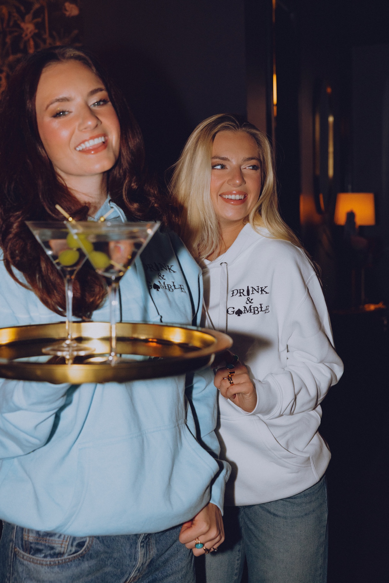 Brunette in a light blue hoodie carrying martinis on a gold tray, blonde in a white hoodie laughing behind