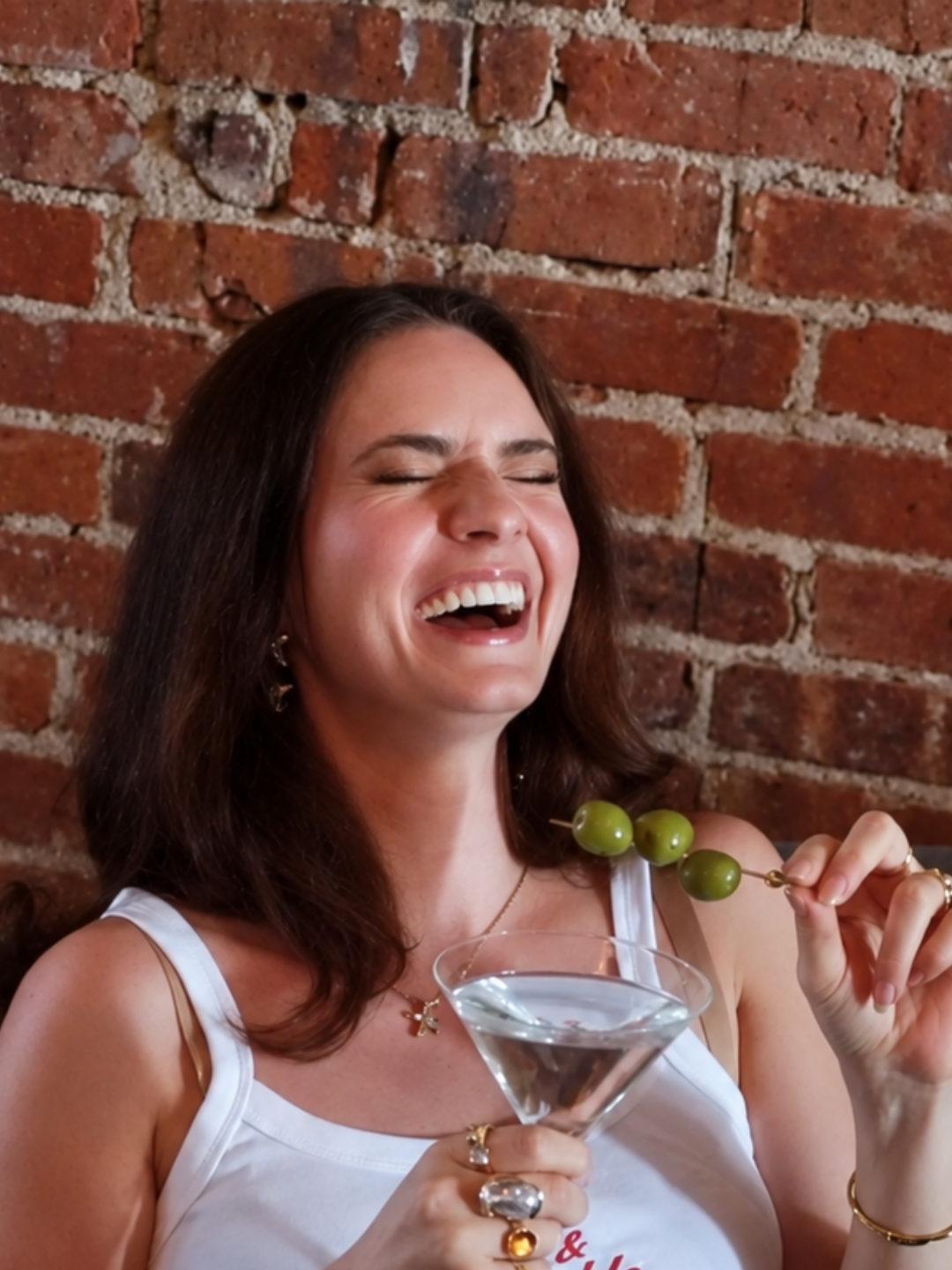 Brunette laughing with a martini and olive skewer against brick