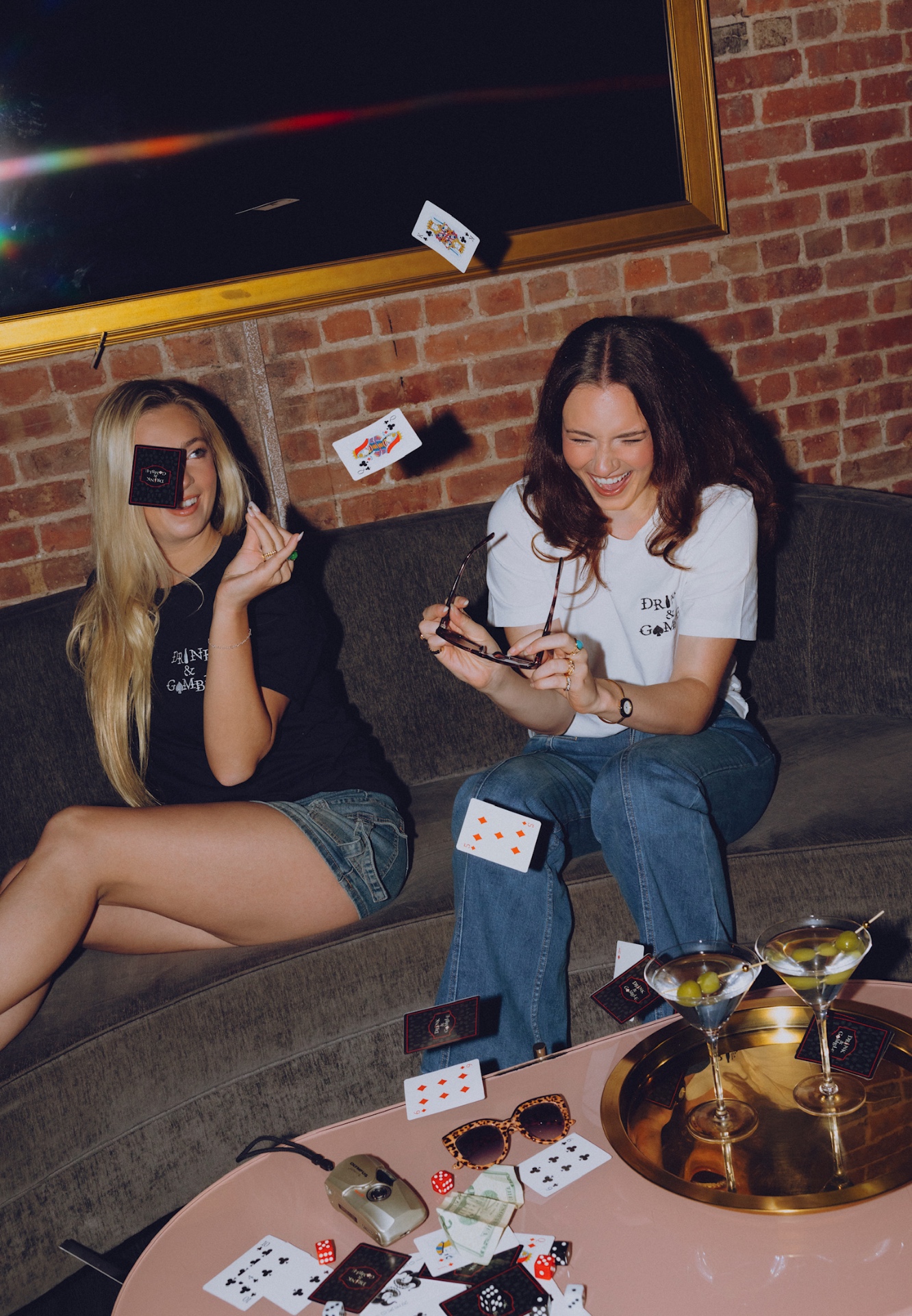 Two women in Drink & Gamble tees throwing playing cards over martinis