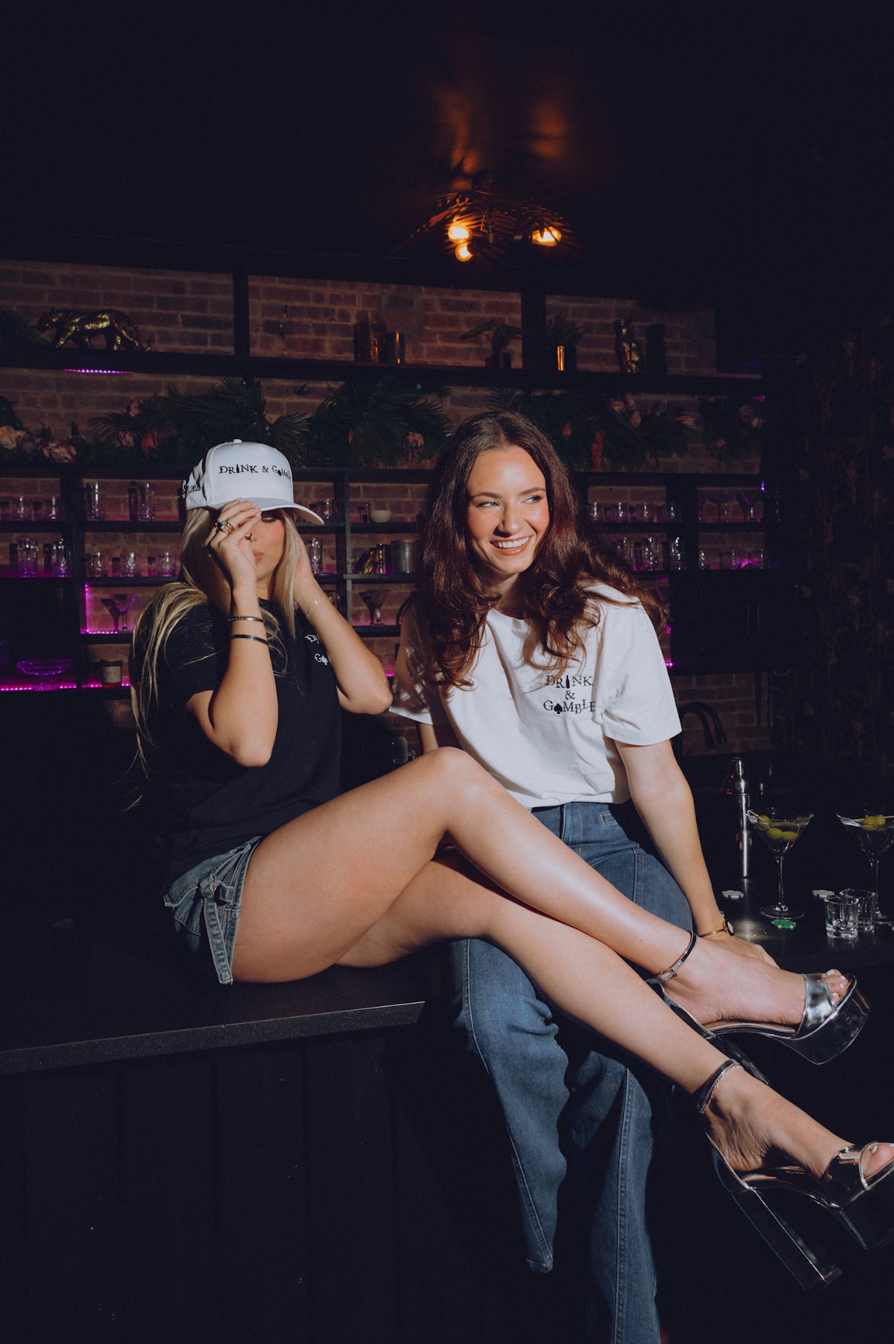 Two women in Drink & Gamble tees and cap sitting on the bar