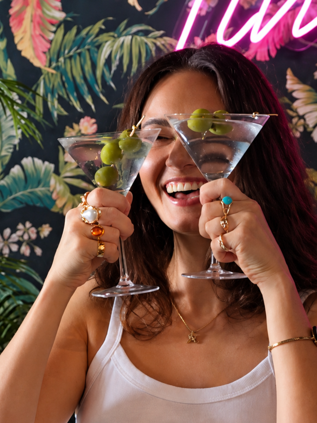 Brunette holding up two martinis in front of tropical wallpaper