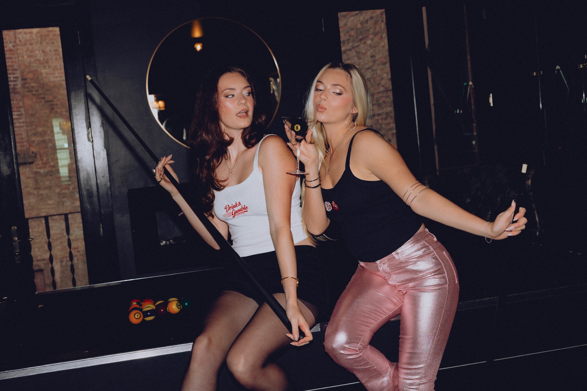 Two women in Drink & Gamble tanks posing with a pool cue and 8-ball martini