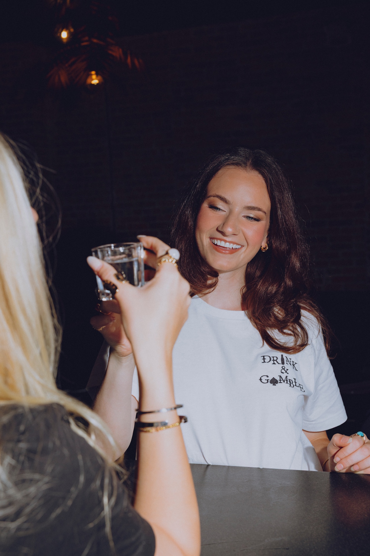 Brunette in a white Drink & Gamble tee toasting at the bar