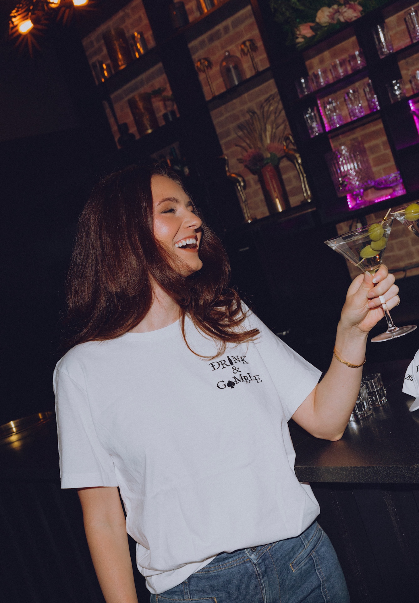 Brunette in a white Drink & Gamble tee laughing with a martini at the bar